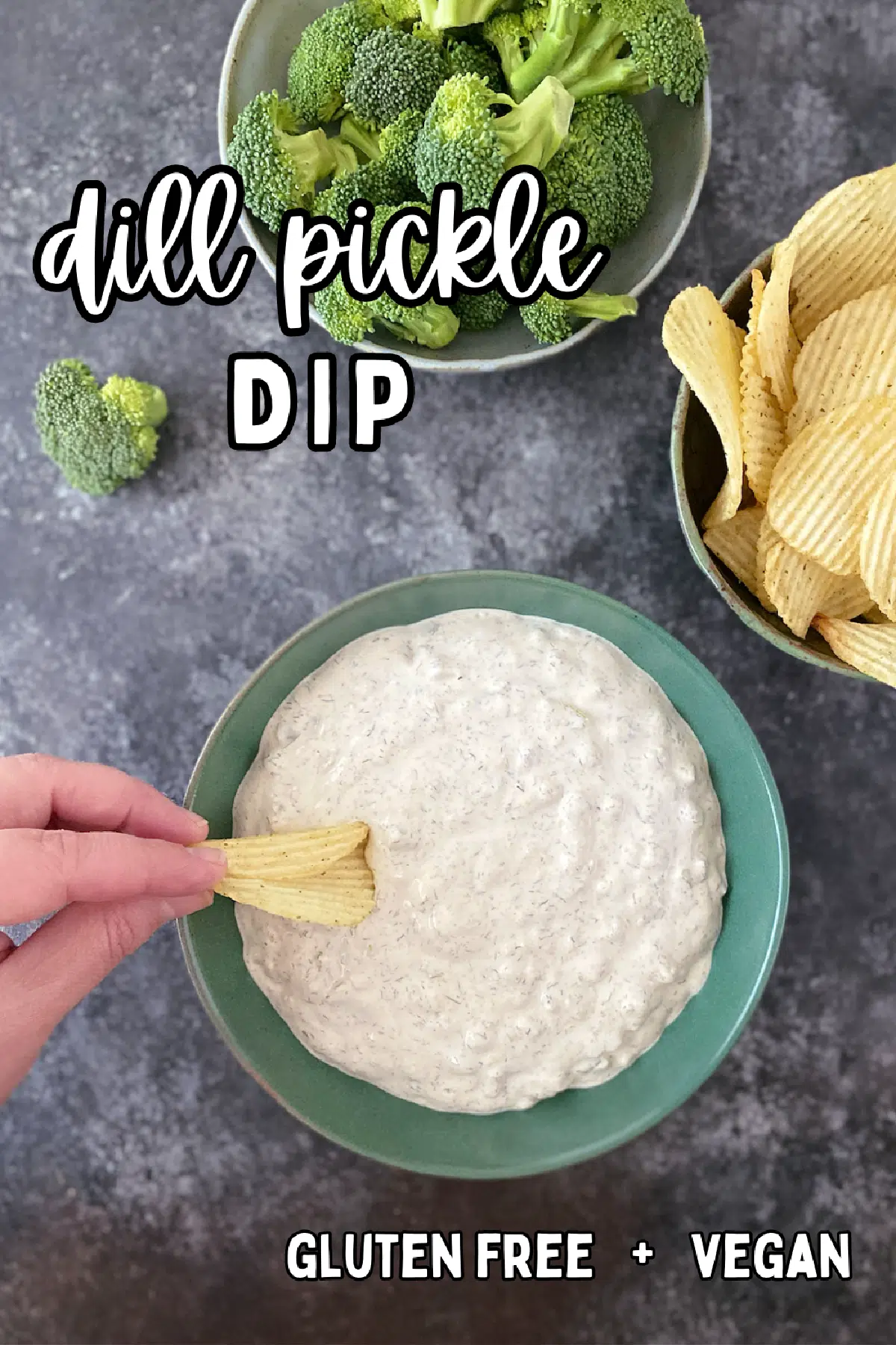 Overhead view of dill pickle dip in a green bowl, with a hand dipping a potato chip in the bowl, next to bowls of ruffled potato chips and broccoli trees for dipping.