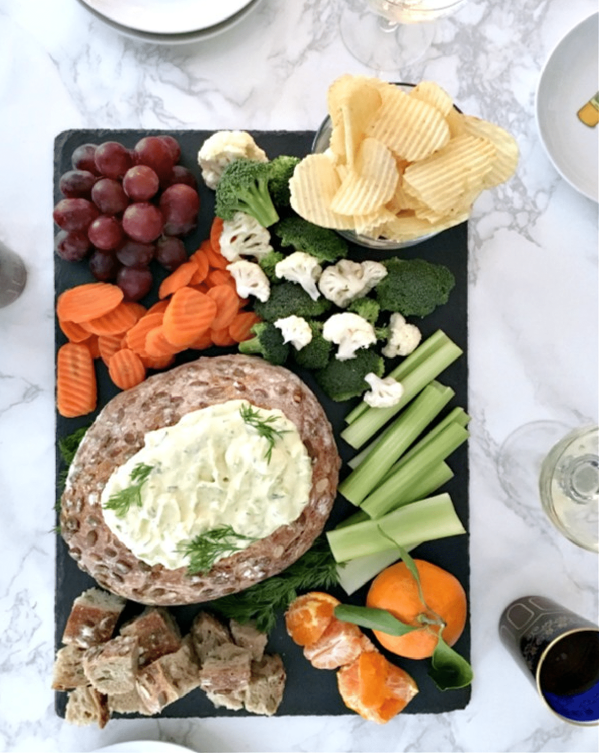 Overhead view of dill pickle dip in a bread bowl, on a black slate snack board with bread cubes, celery sticks, broccoli and cauliflower trees, ruffled potato chips, a bunch of grapes, and sliced carrots.