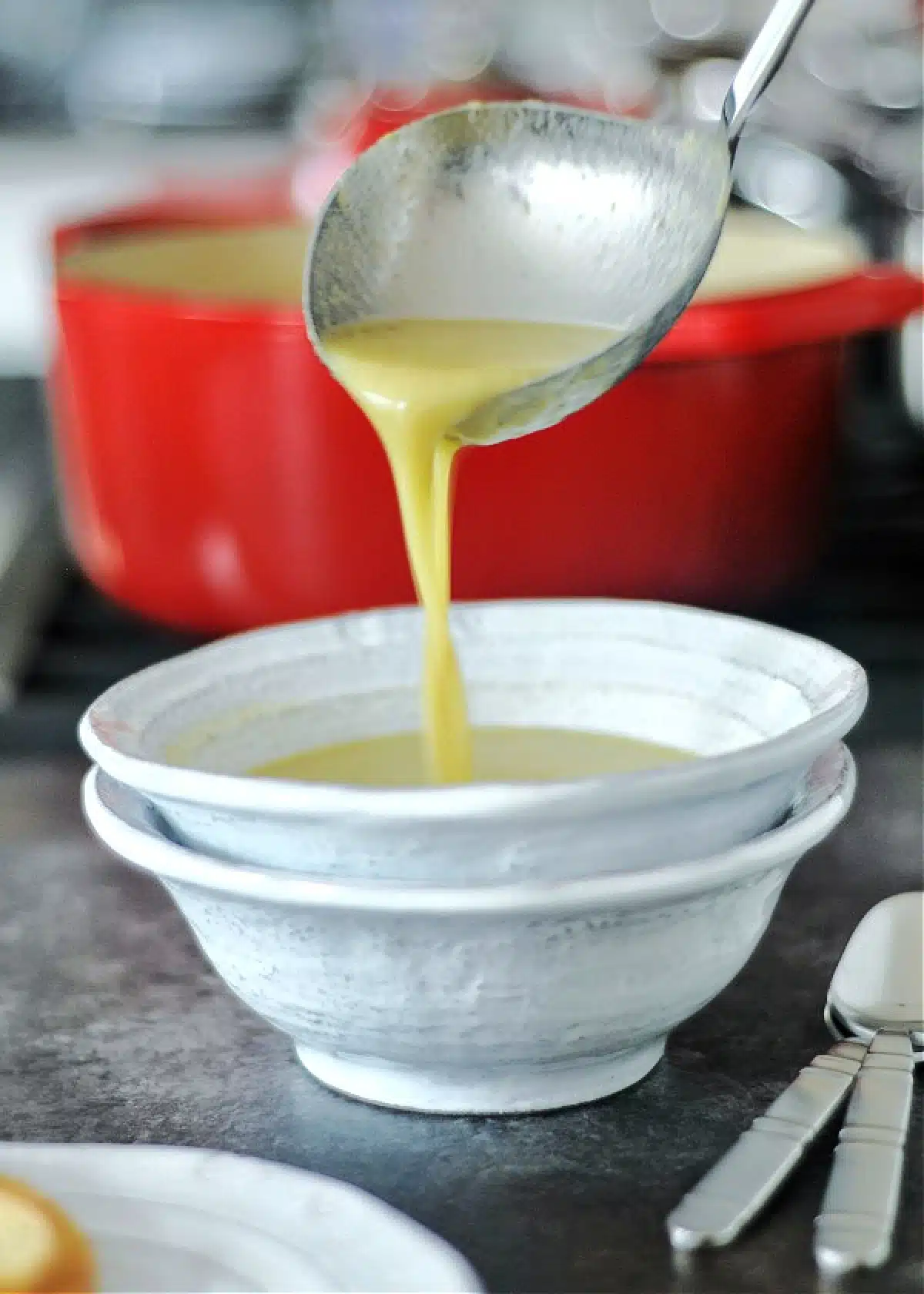 Silver ladle pouring artichoke soup into a bowl, large red soup pot blurred in background.