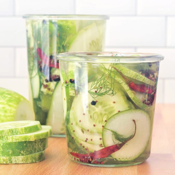 Two glass jars of quick pickles sit side by side on a butcher block, with a small stack of raw sliced cucumber sitting alongside.