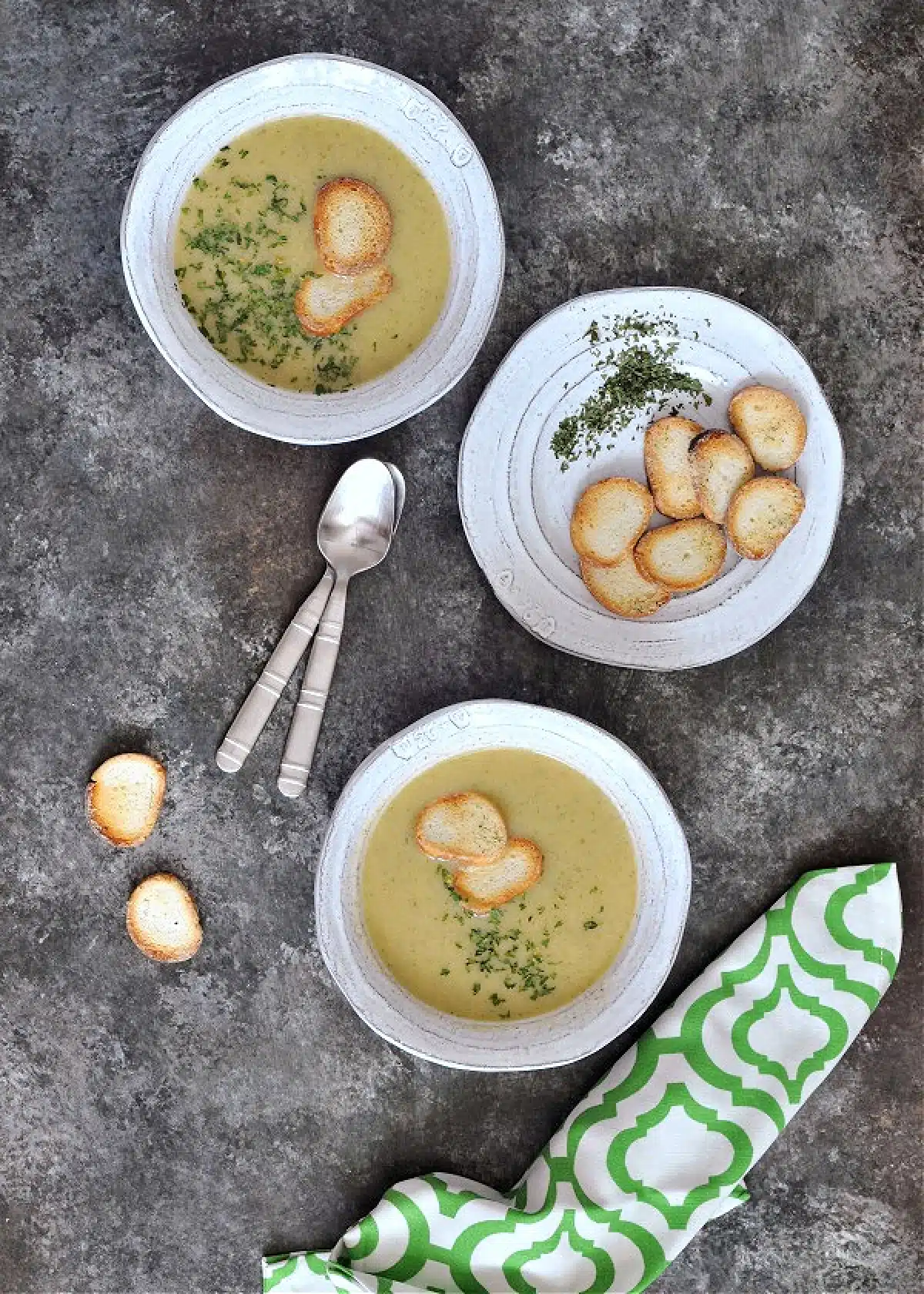 Overhead view of artichoke soup in bowls garnished with crisp bagel chips and chopped herbs.