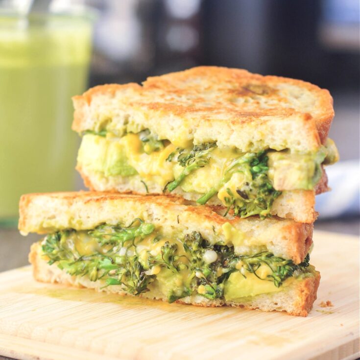 Green Goddess grilled cheese sandwich sliced in half and stacked on a small light colored wood cutting board. The sandwich has Green Goddess dressing, broccolini trees, sliced avocado, and melted dairy free cheese. A glass jar of Green Goddess dressing is blurred in the background.