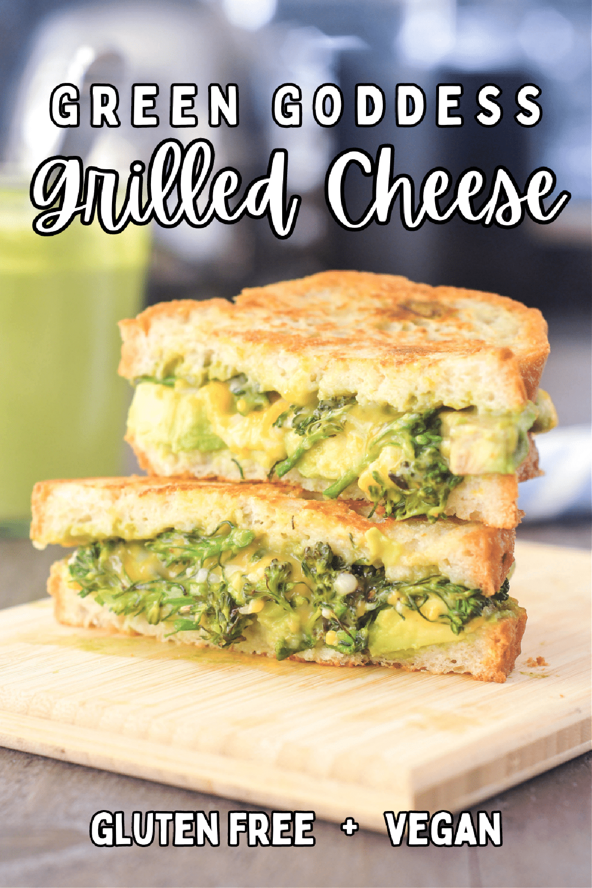 Green Goddess grilled cheese sandwich sliced in half and stacked on a small light colored wood cutting board. The sandwich has Green Goddess dressing, broccolini trees, sliced avocado, and melted dairy free cheese. A glass jar of Green Goddess dressing is blurred in the background.