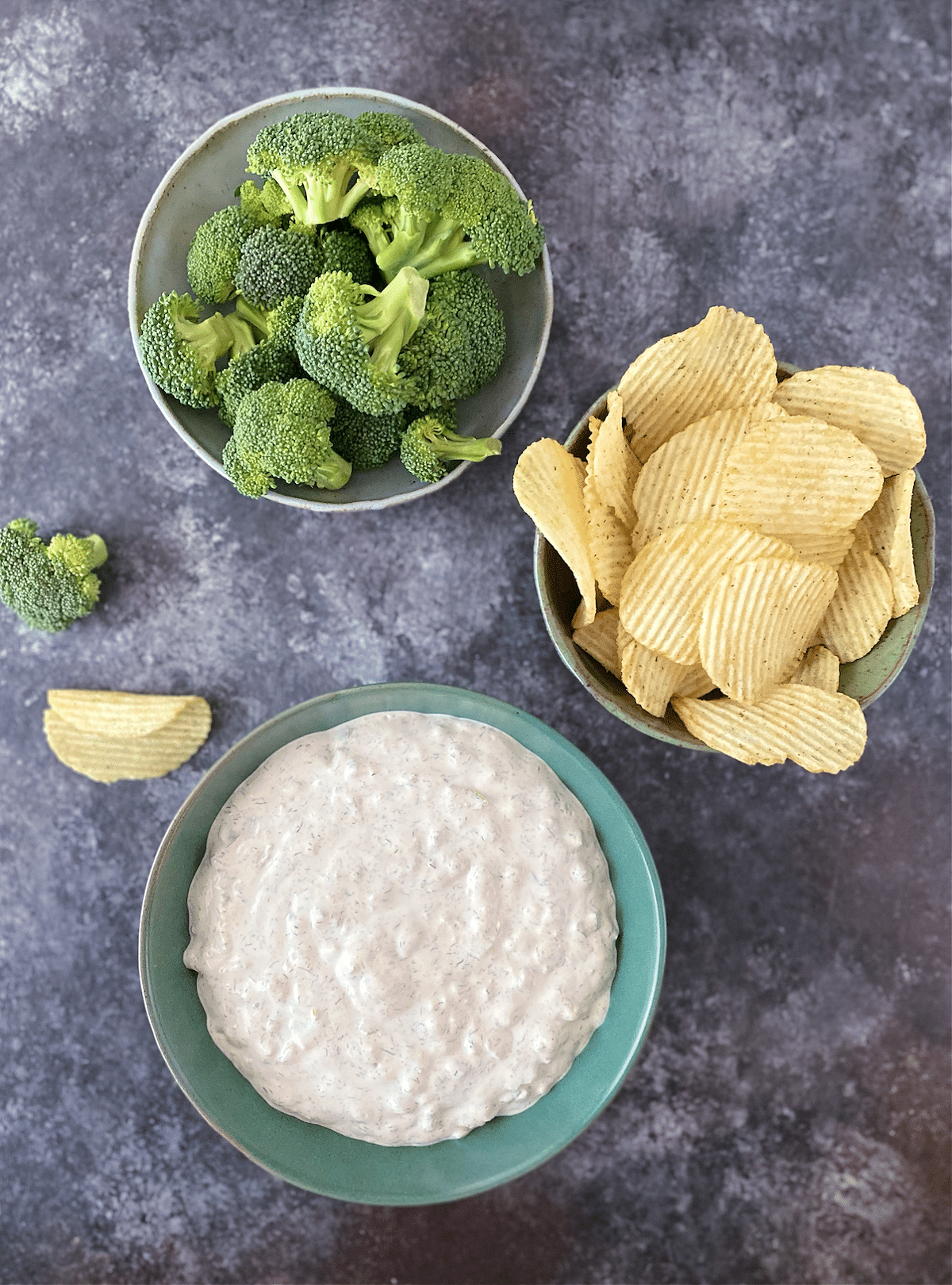 Overhead view of dill pickle dip in a green bowl, with bowls of ruffled potato chips and broccoli trees for dipping.