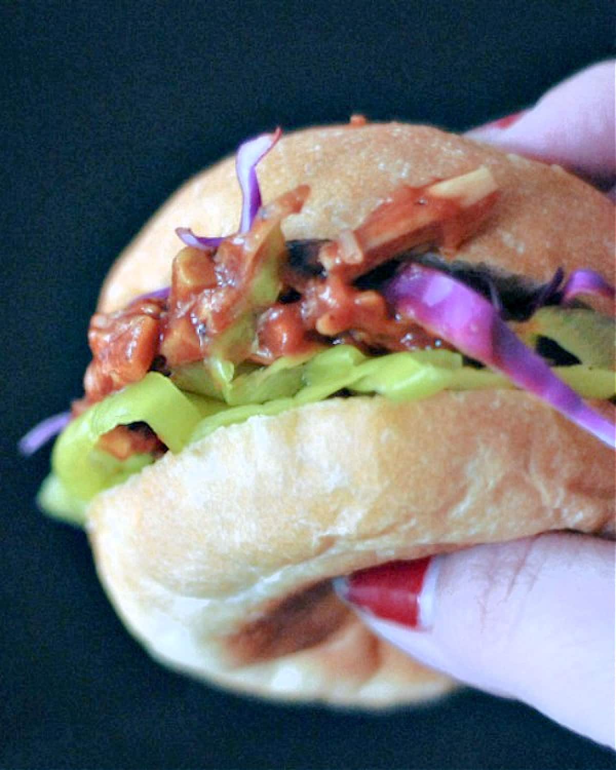 A hand holding a vegan pulled porcini slider with purple cabbage and pepperoncini pepper rings.