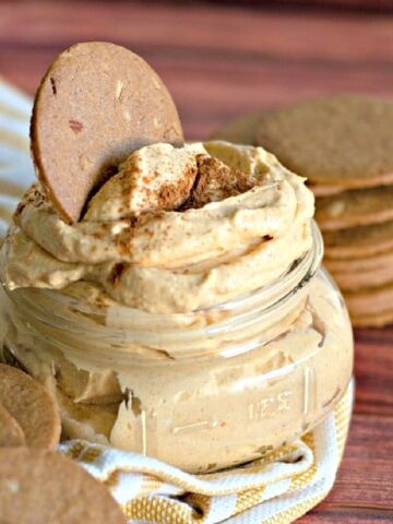 A ginger cookie dipped into a glass jar of pumpkin cheesecake dip. Jar sits on a yellow and white striped cloth napkin, stack of thin crispy ginger cookies next to napkin.
