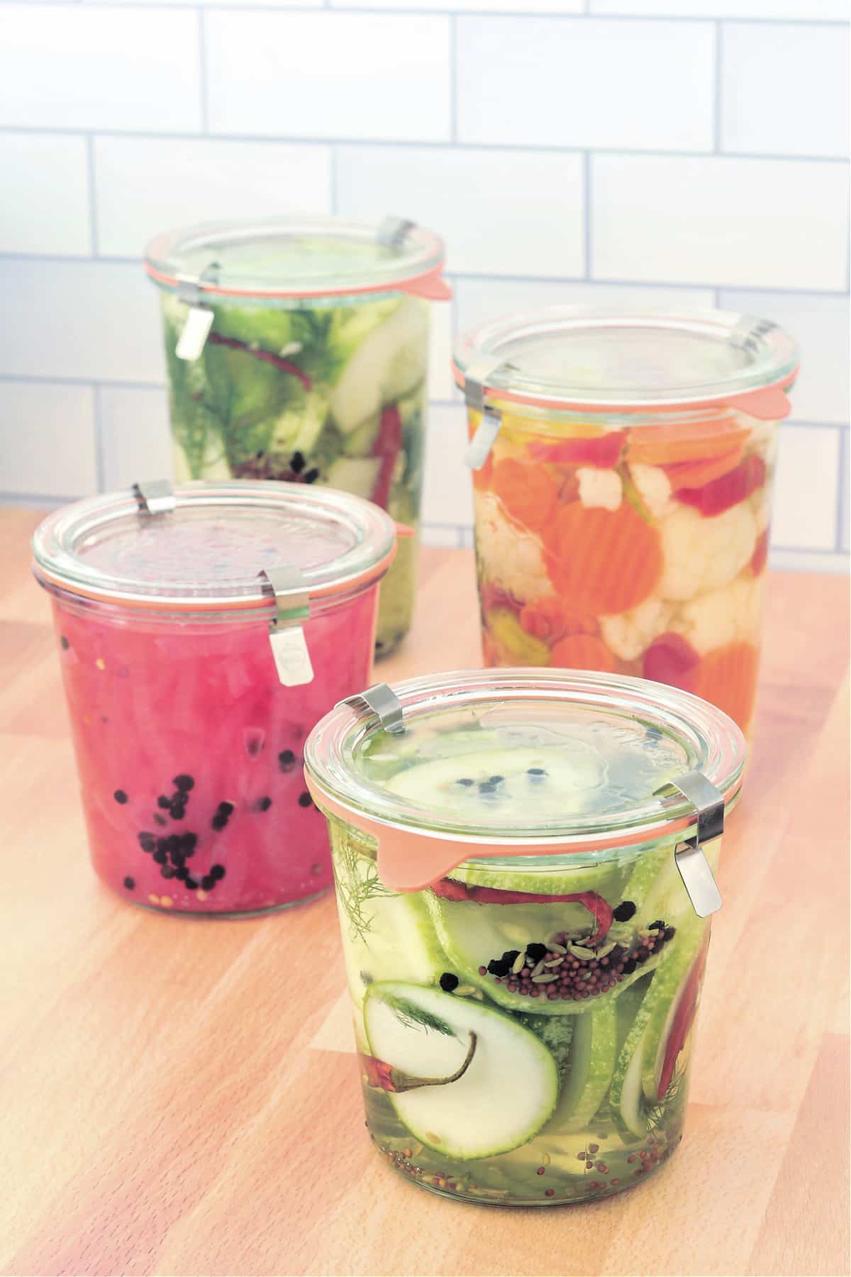 Four glass jars filled with quick pickles sit on a butcher block: a small jar of bright pink pickled onions sits on a larger jar of quick pickles, and a smaller jar of quick pickles sits on a larger jar of pickled carrot, cauliflower, and peppers.