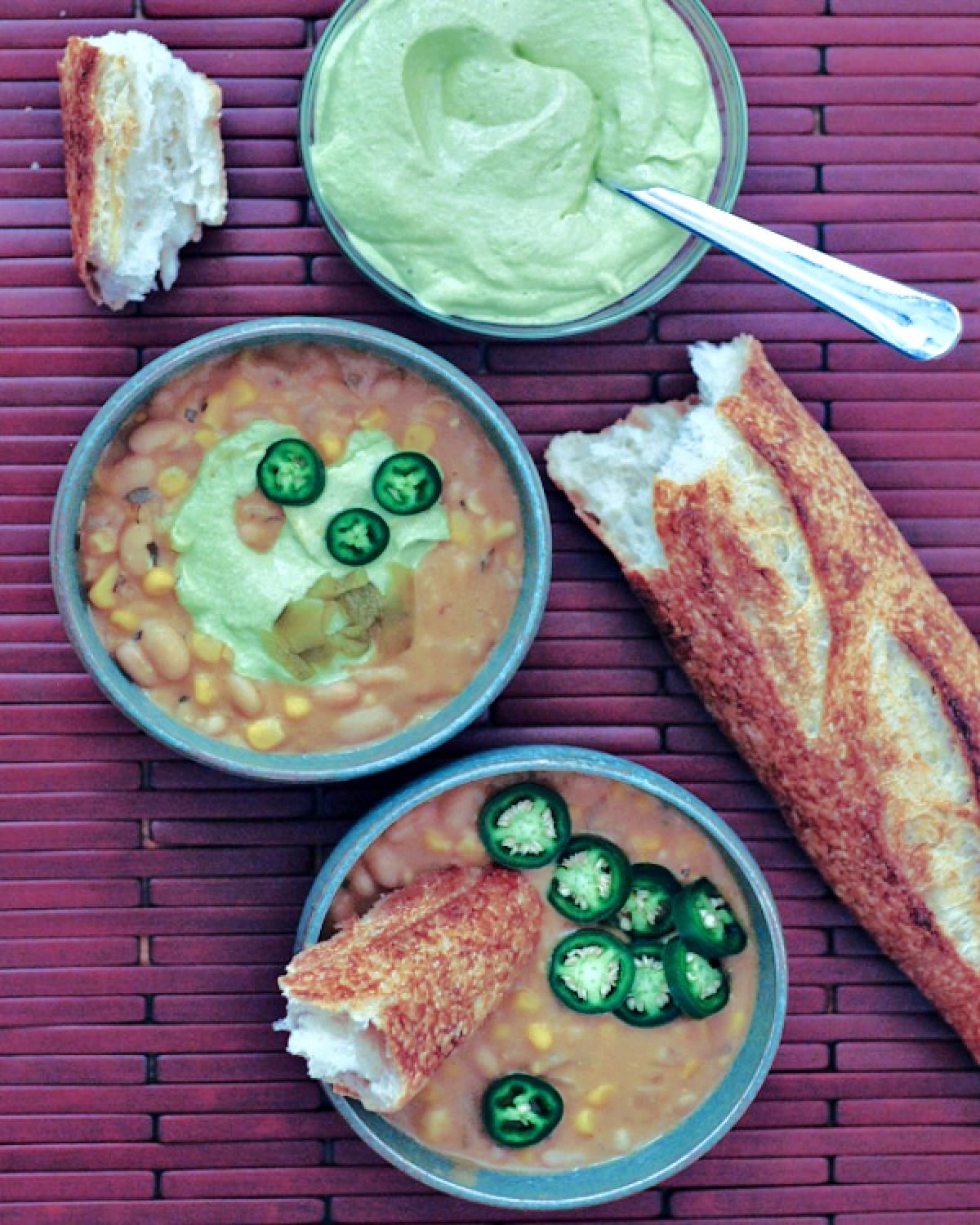 Overhead view of white bean chili in grey bowls, garnished with light green Hatch avocado cream, a piece of crusty baguette, sliced jalapeno peppers.