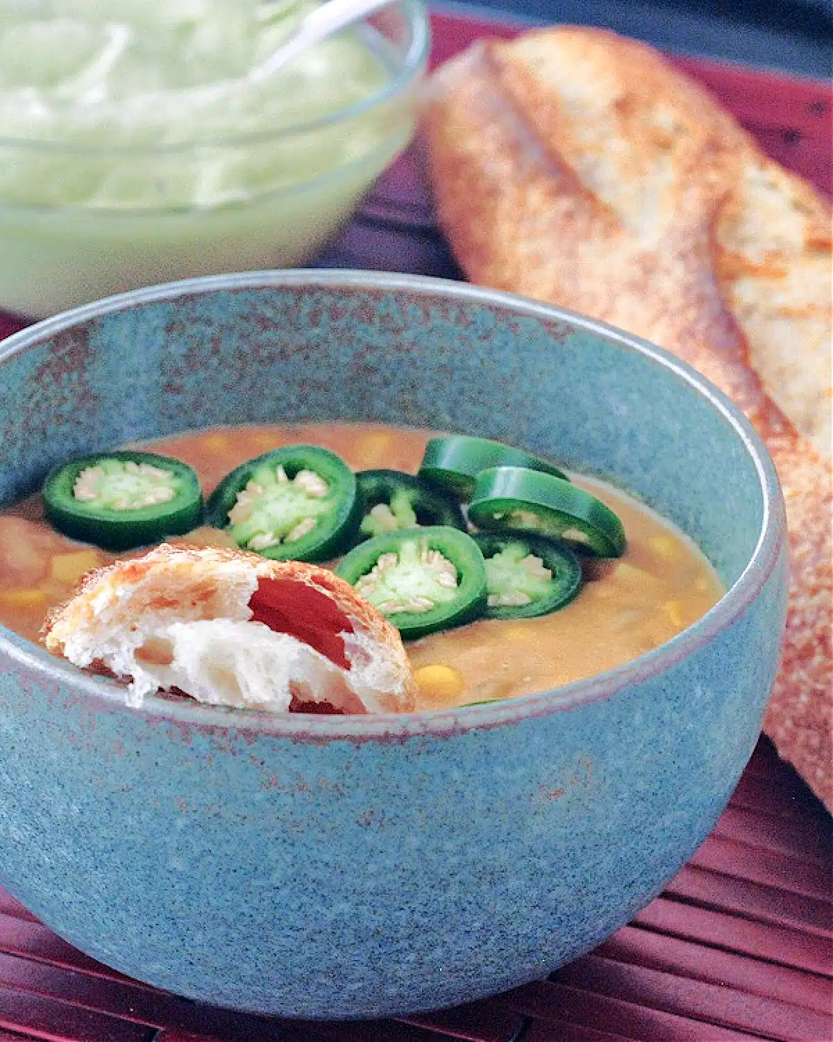 White bean chili in a grey bowl, garnished with light green Hatch avocado cream, a piece of crusty baguette, sliced jalapeno peppers.