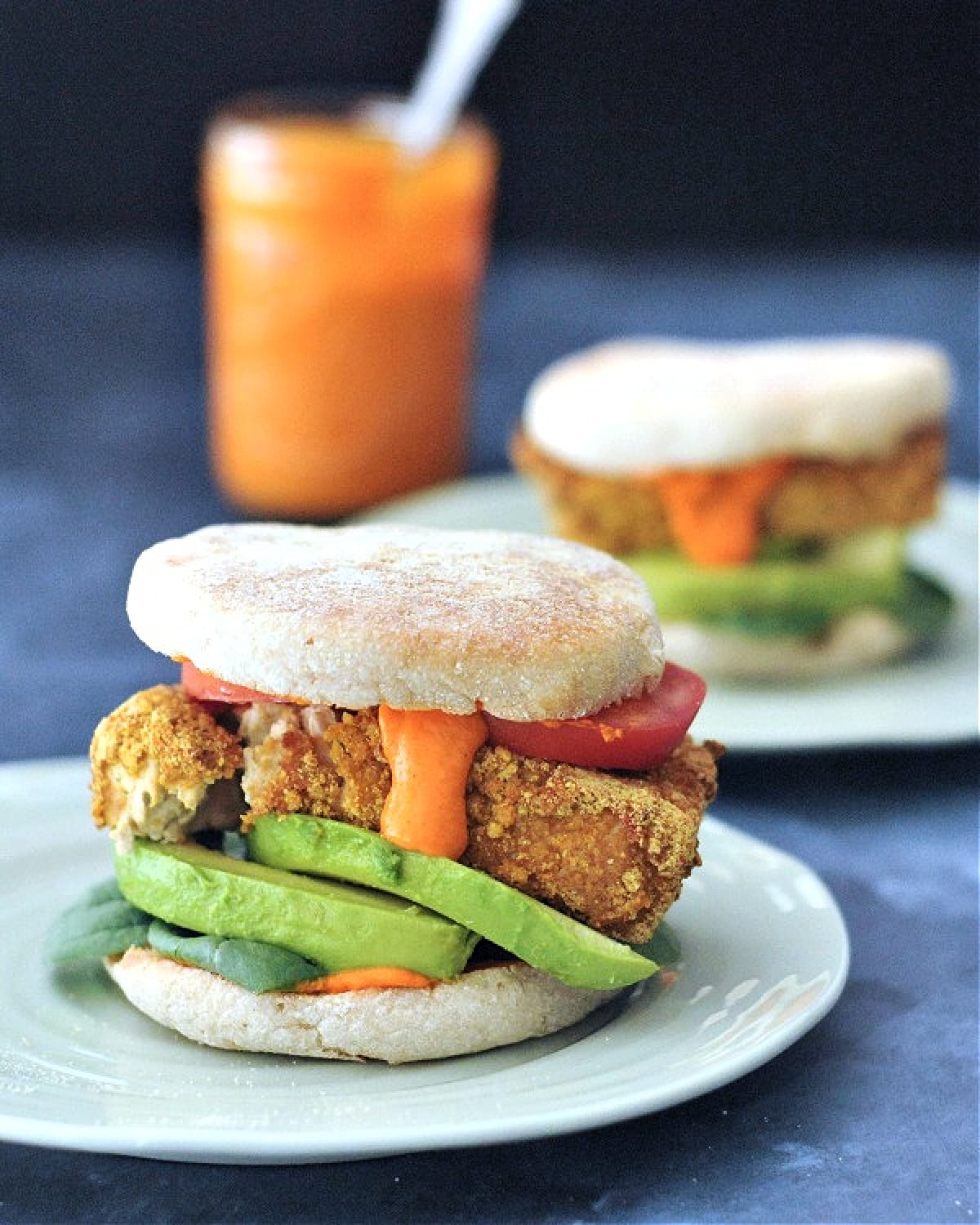 Two vegan breakfast sandwiches on white plates, side view shows stacked ingredients (Romesco sauce, spinach, tomato slices, vegan "egg" or tofu, avocado slices).