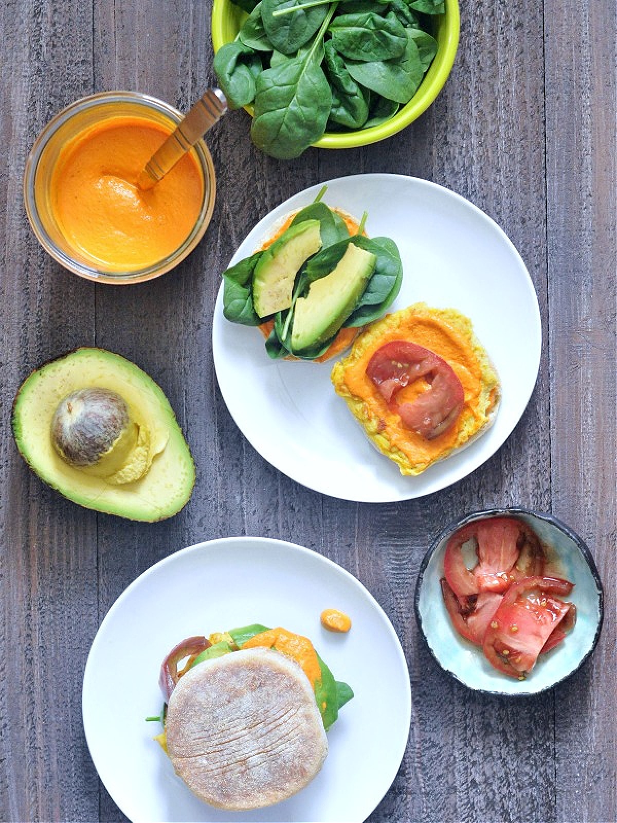 Overhead view of two vegan breakfast sandwiches on white plates, one open faced to show ingredients (Romesco sauce, spinach, tomato slices, vegan "egg" or tofu, avocado slices).