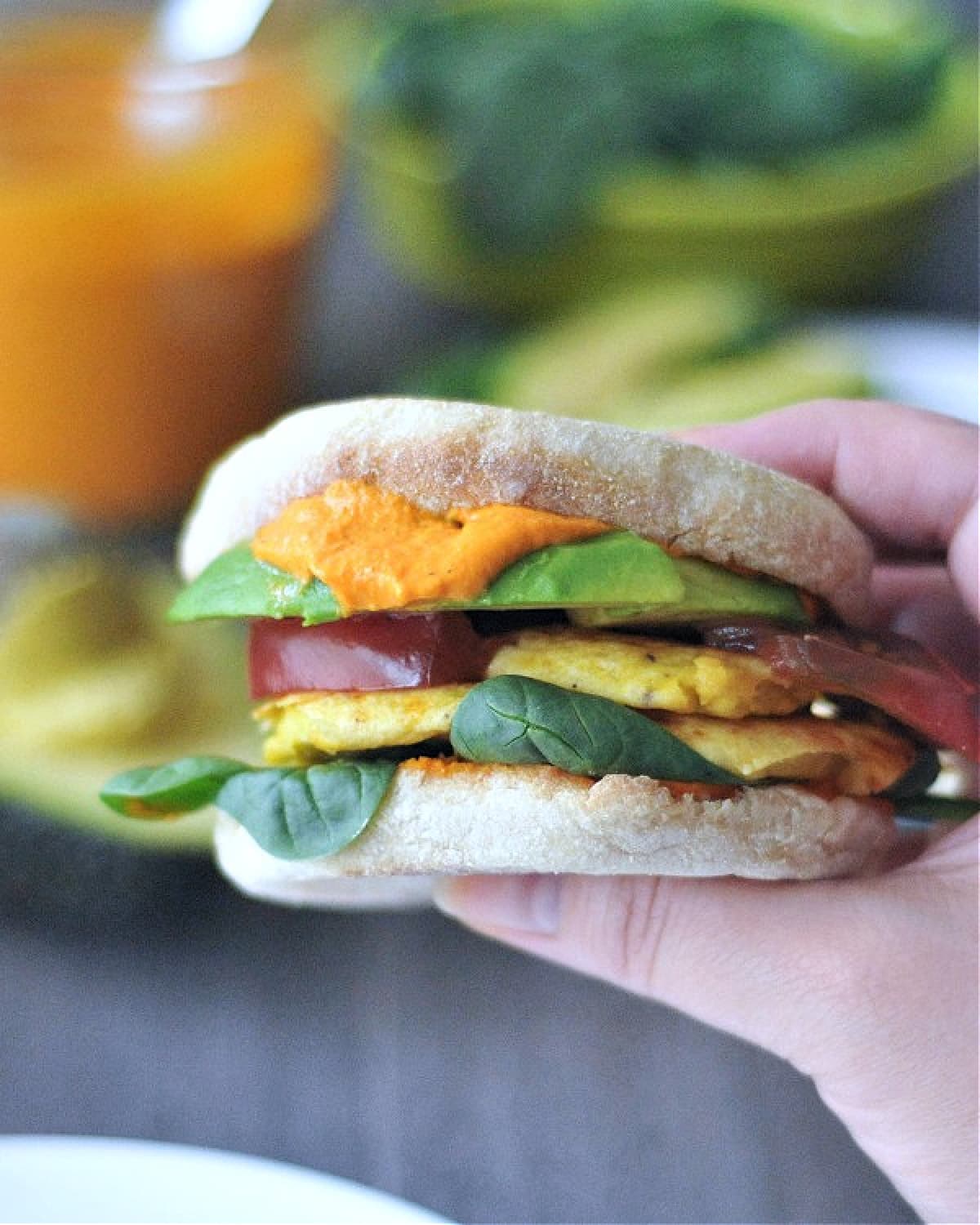 A hand holding a vegan breakfast sandwich, side view shows stacked ingredients (Romesco sauce, spinach, tomato slices, vegan "egg" or tofu, avocado slices).