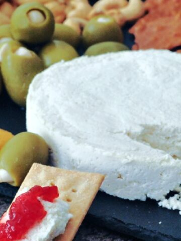 A wheel of vegan brie cheese on a slate board with crackers, olives, nuts, and veggies.