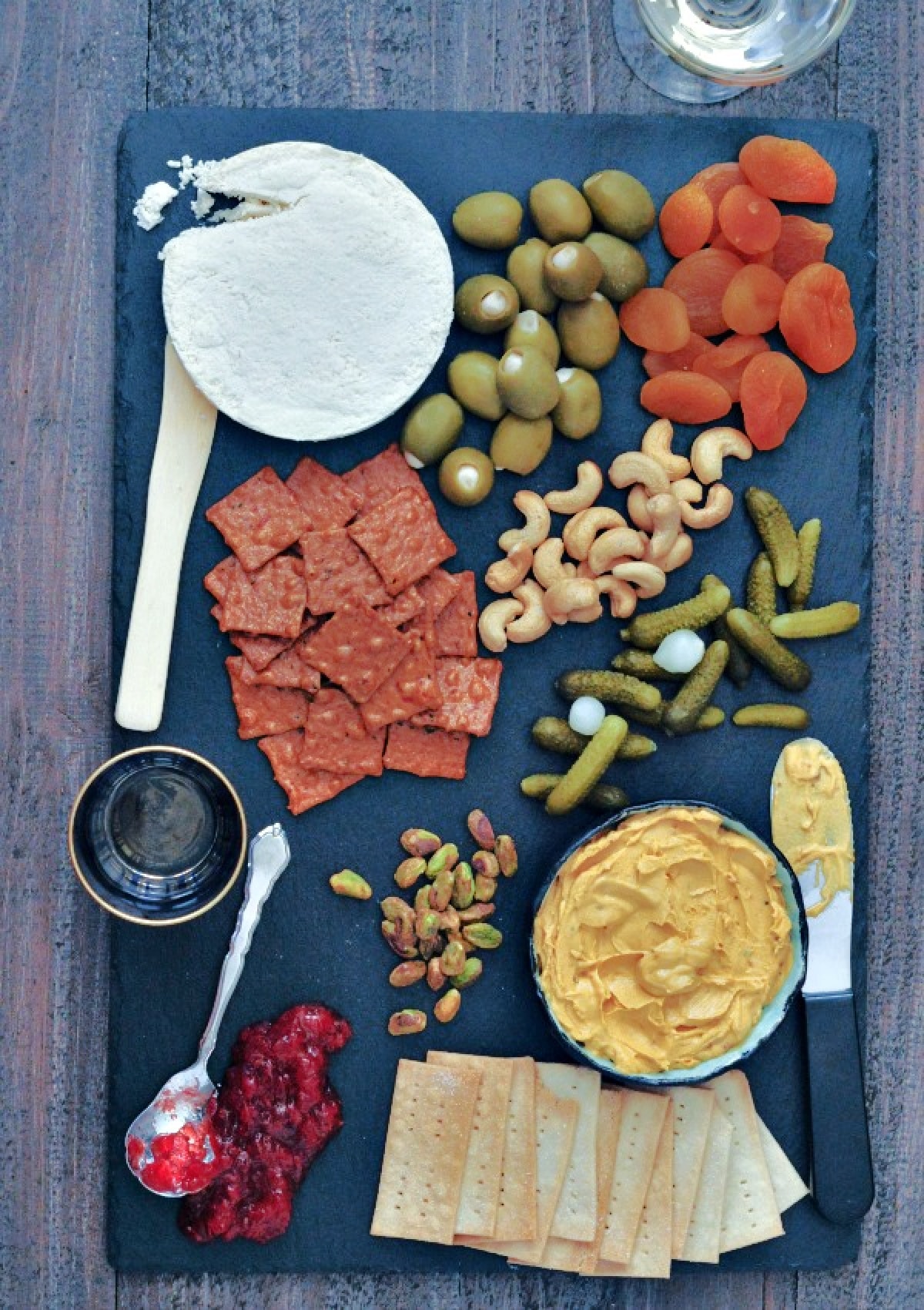 Overhead view of a wheel of vegan brie cheese on a slate board with crackers, olives, nuts, and veggies.