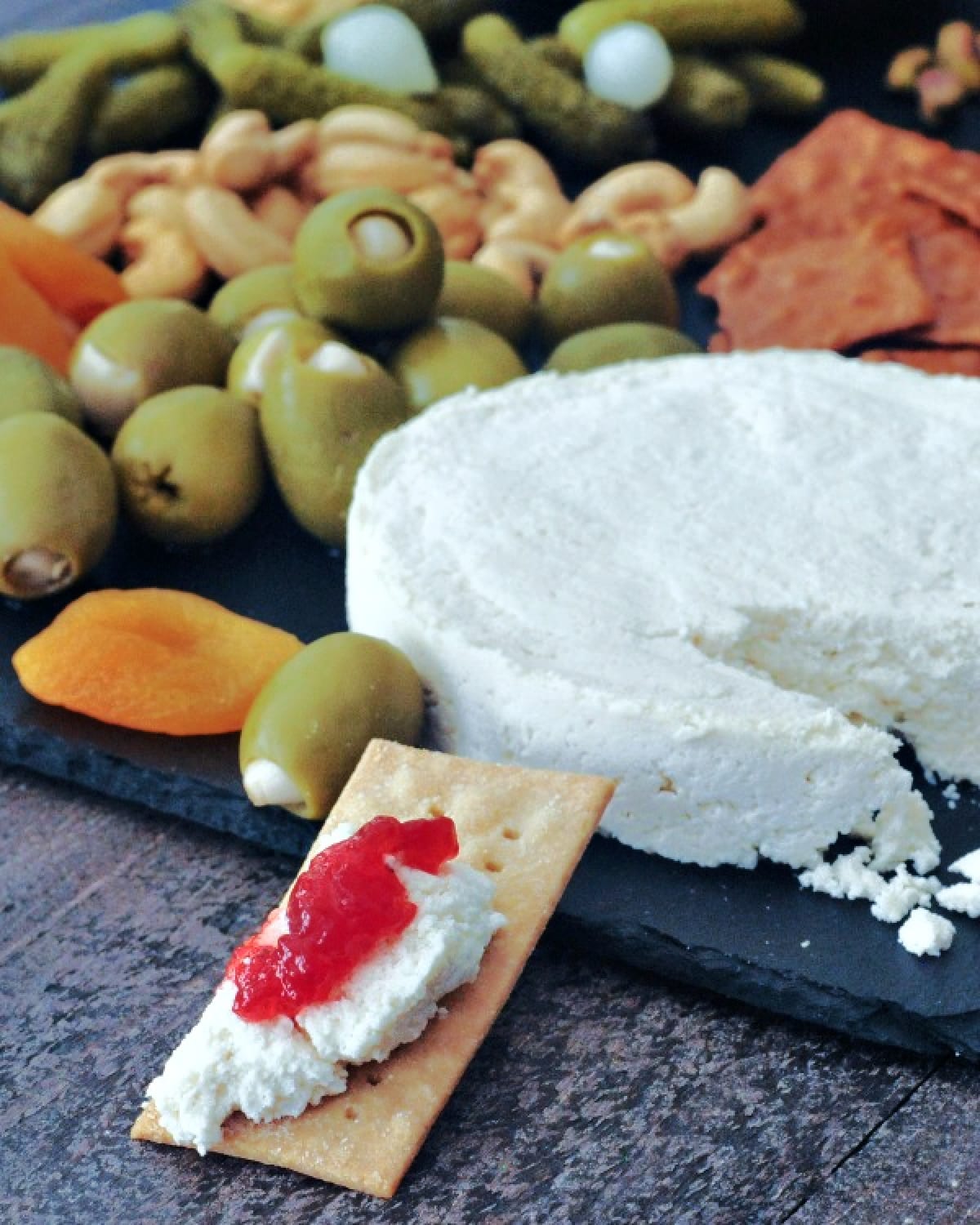 A wheel of vegan brie cheese on a slate board with crackers, olives, nuts, and veggies. A cracker spread with vegan brie and bright red fruit jam sits in front of the cheese board.