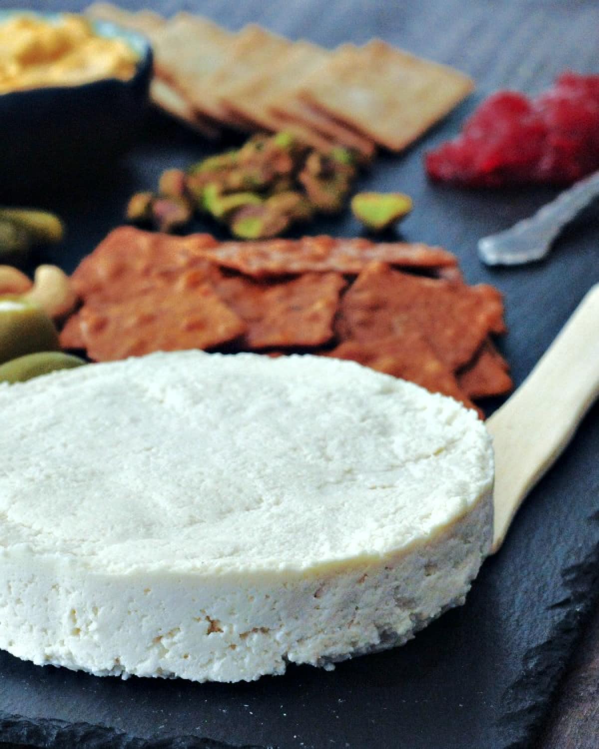 A wheel of vegan brie cheese on a slate board with crackers, olives, nuts, and veggies.