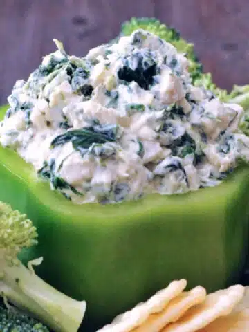 Spinach dip served in a hollowed out green bell pepper, with fresh broccoli trees and rice crackers for dipping.