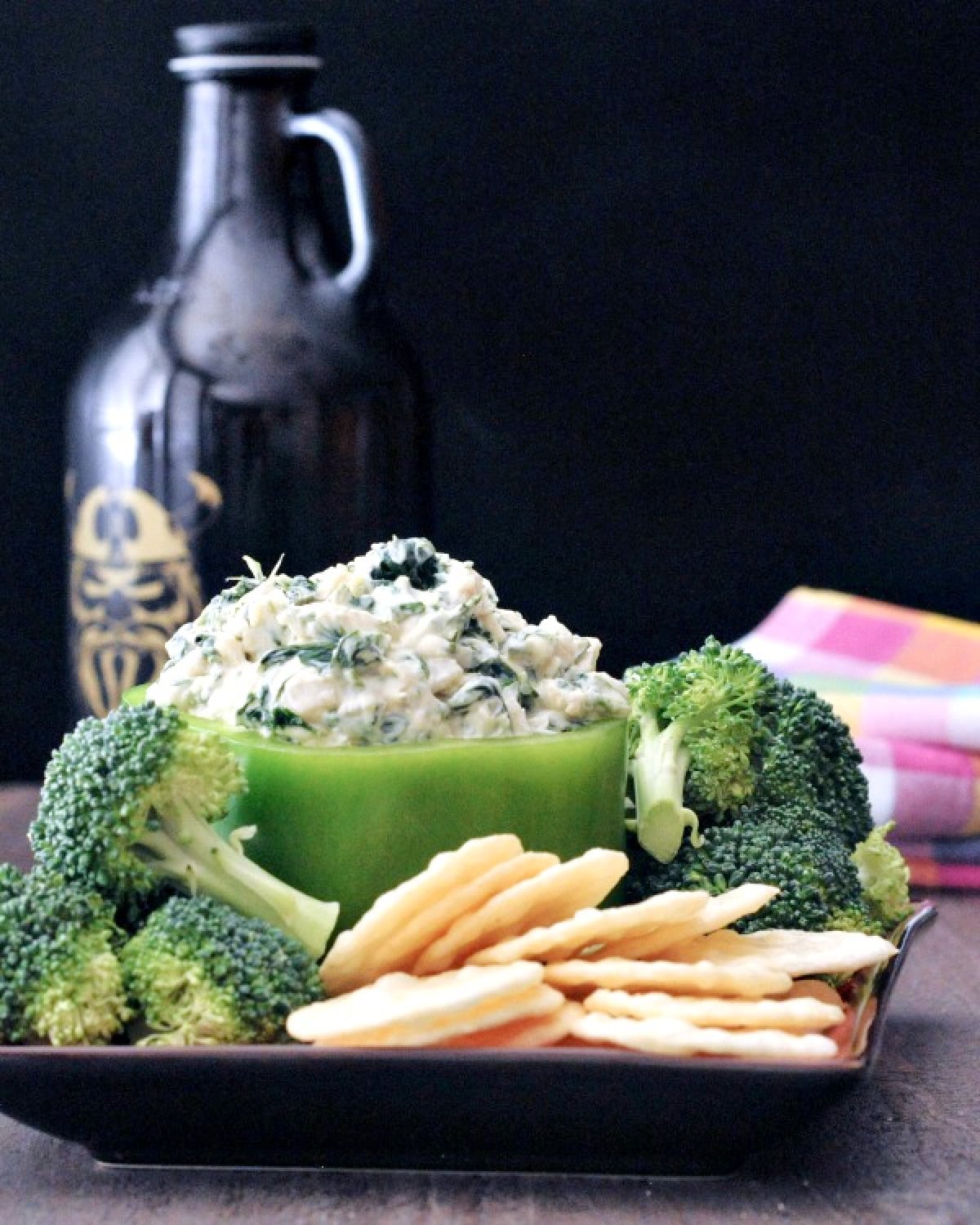 Spinach dip served in a hollowed out green bell pepper, with fresh broccoli trees and rice crackers for dipping.