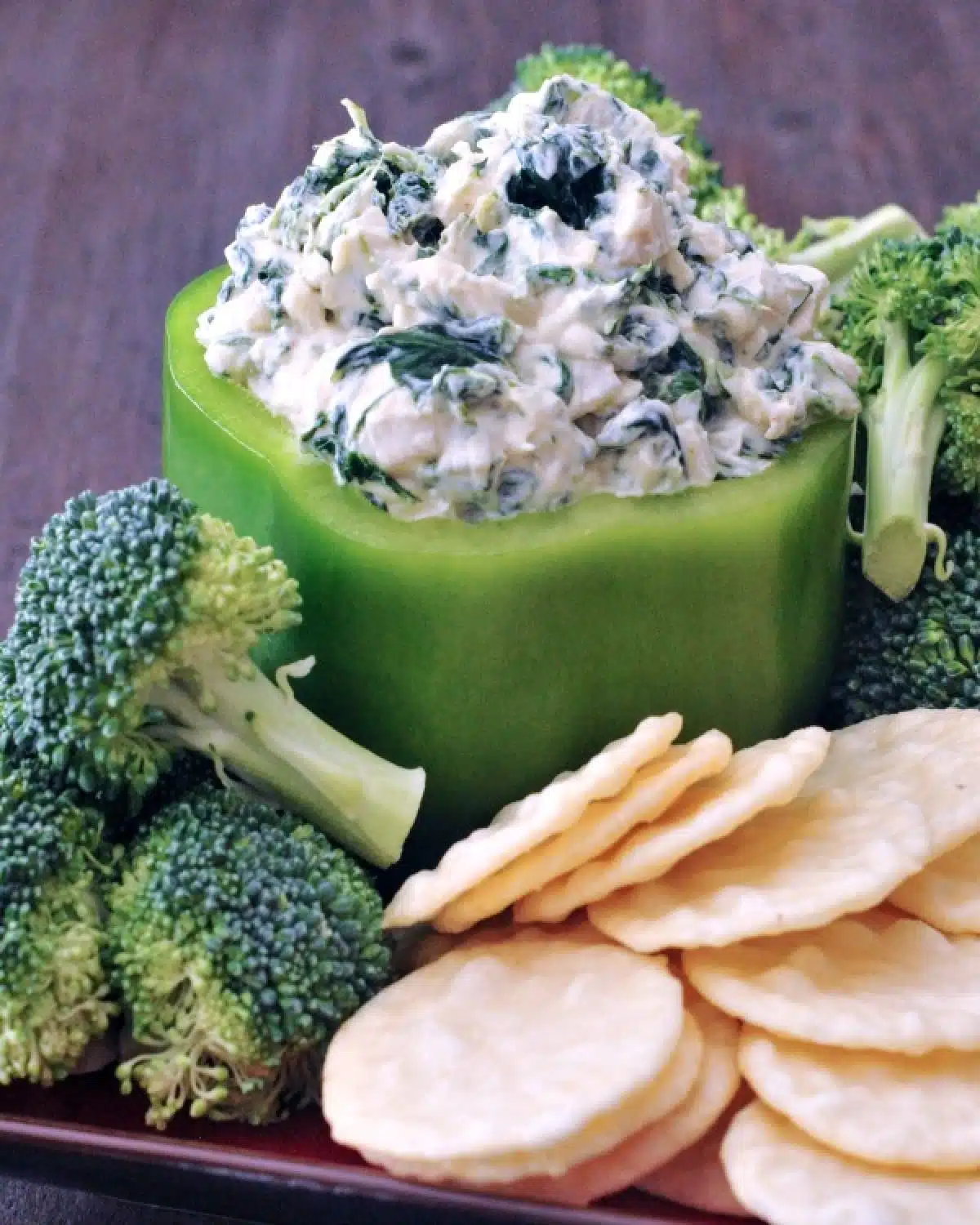 Spinach dip served in a hollowed out green bell pepper, with fresh broccoli trees and rice crackers for dipping.