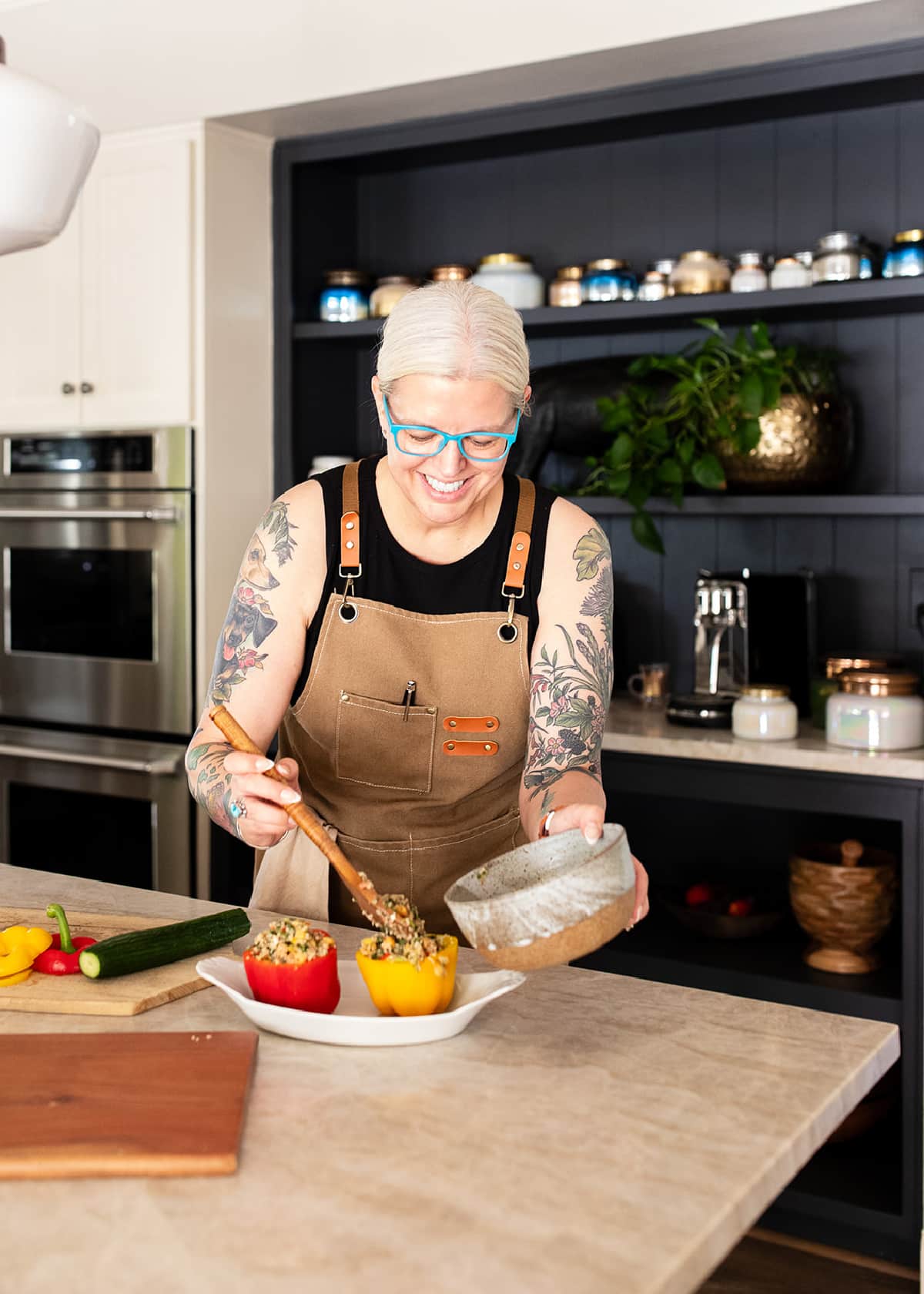 Kristina standing at a counter in a sleeveless black top and a tan chefs apron, holding a bowl of quinoa salad as she spoons the salad into a hollowed out bell pepper.