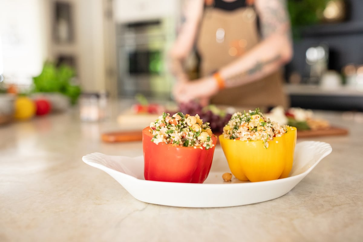 Two quinoa salad stuffed bell peppers (one red, one yellow pepper) sit in a white oval dish, while blurred in the background, Kristina is chopping vegetables.