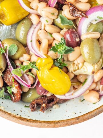 Close up overhead view of a bowl of white bean salad, which includes black kalamata and green castelvetrano olives, white beans, sun dried tomatoes, sliced artichoke hearts, sliced red onion, pepperoncini peppers, herbs and spices.