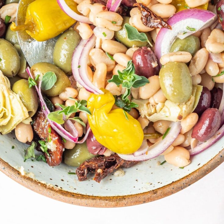 Close up overhead view of a bowl of white bean salad, which includes black kalamata and green castelvetrano olives, white beans, sun dried tomatoes, sliced artichoke hearts, sliced red onion, pepperoncini peppers, herbs and spices.