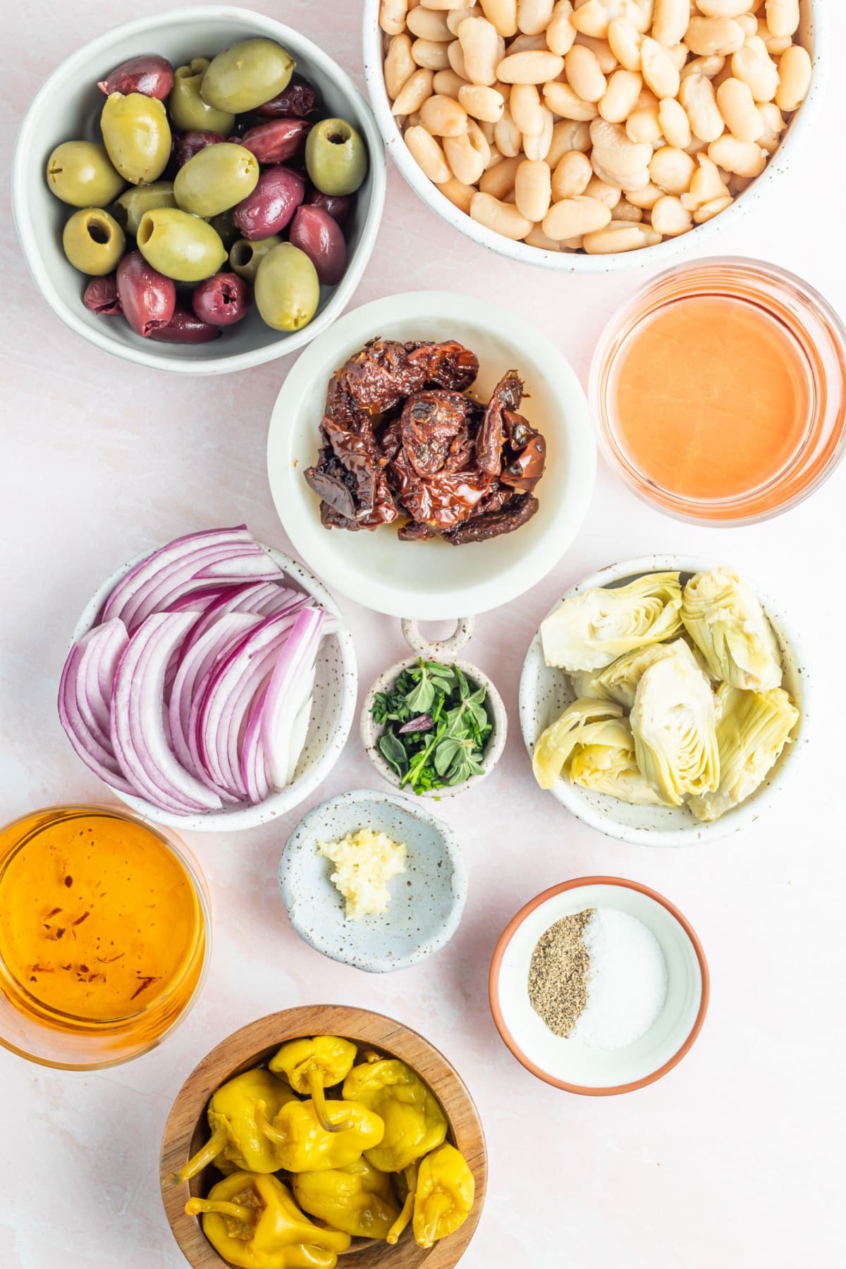 Overhead view of separate bowls of ingredients for a white bean antipasto salad, including black kalamata and green castelvetrano olives, white beans, sun dried tomatoes, sliced artichoke hearts, sliced red onion, pepperoncini peppers, herbs and spices.