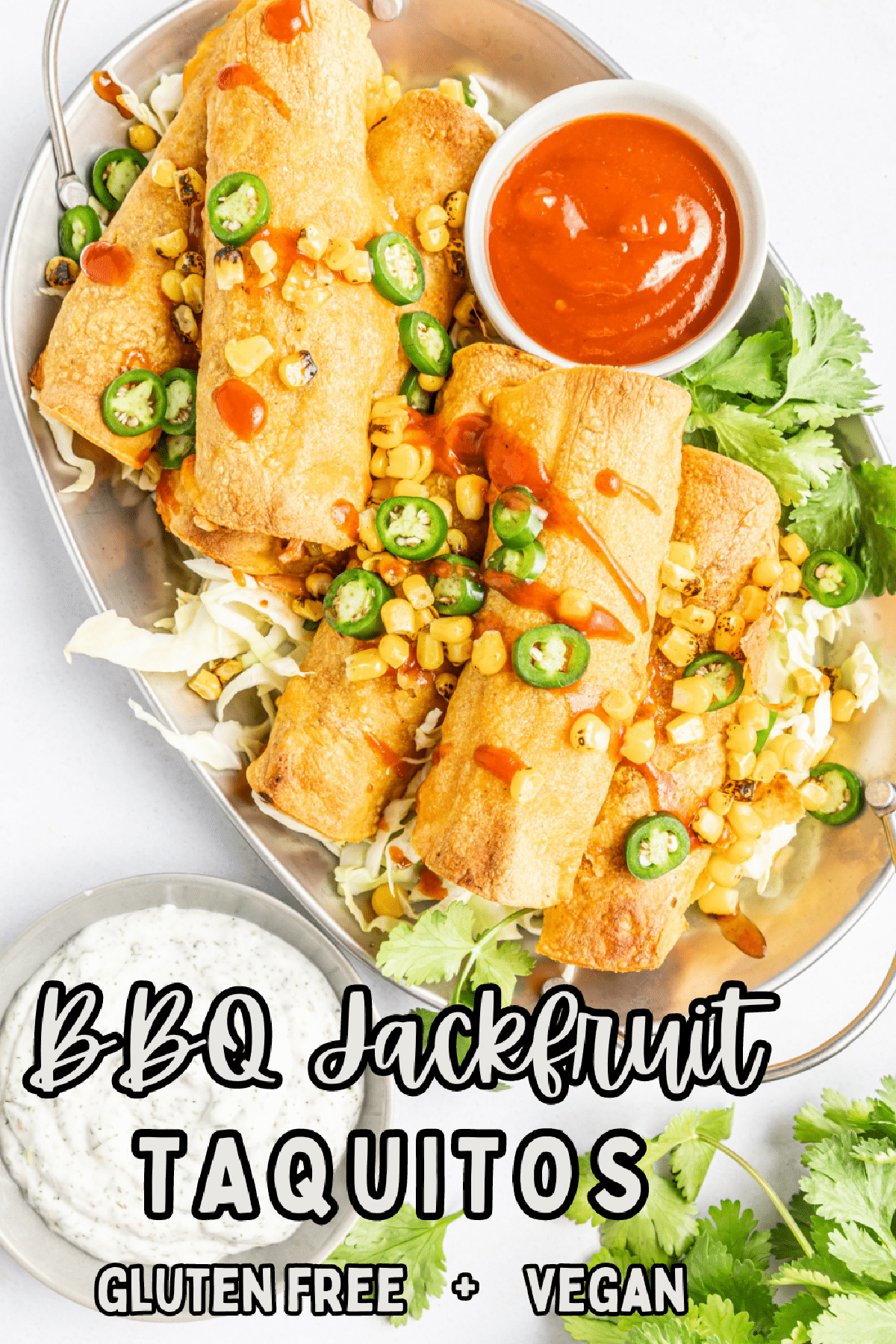Overhead view of BBQ jackfruit taquitos piled onto a silver platter, topped with fire roasted corn, jalapeno slices, cilantro, and a drizzle of BBQ sauce. Small bowls of BBQ sauce and ranch dressing on the side.