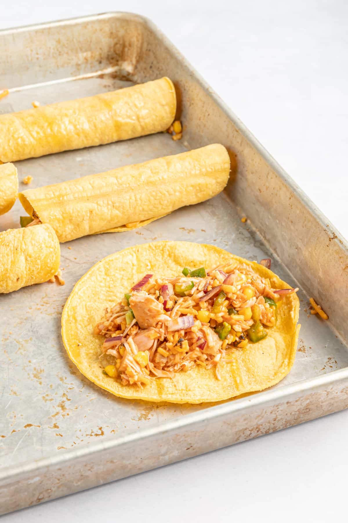 A baking sheet of filled and rolled taquitos, one tortilla laying open flat to show filling inside.