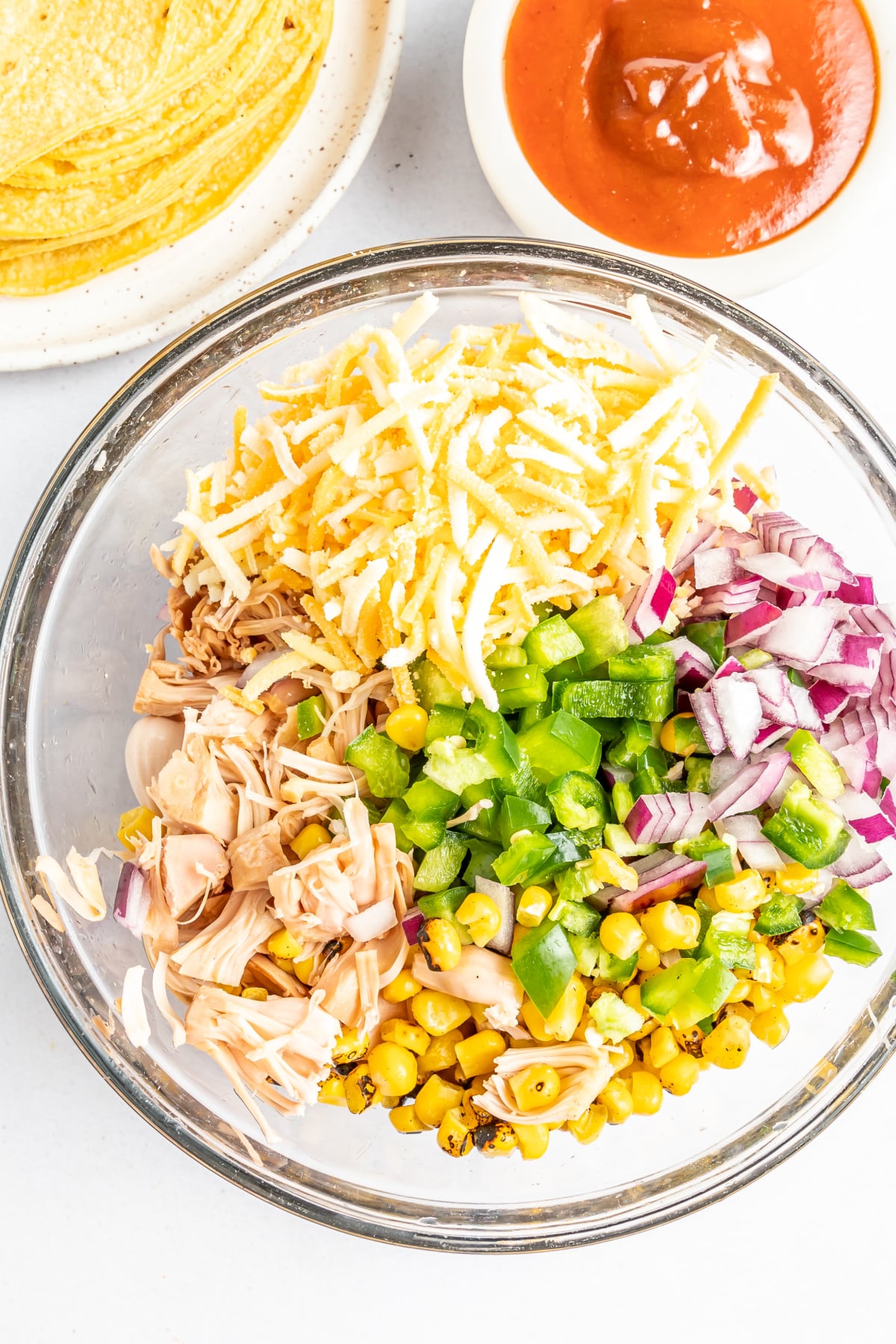 Large mixing bowl of ingredients for BBQ taquitos before mixing: jackfruit, cheese, fire roasted corn, died red onion, and diced jalapeno peppers. A smaller bowl of BBQ sauce and a stack of corn tortillas on the side.
