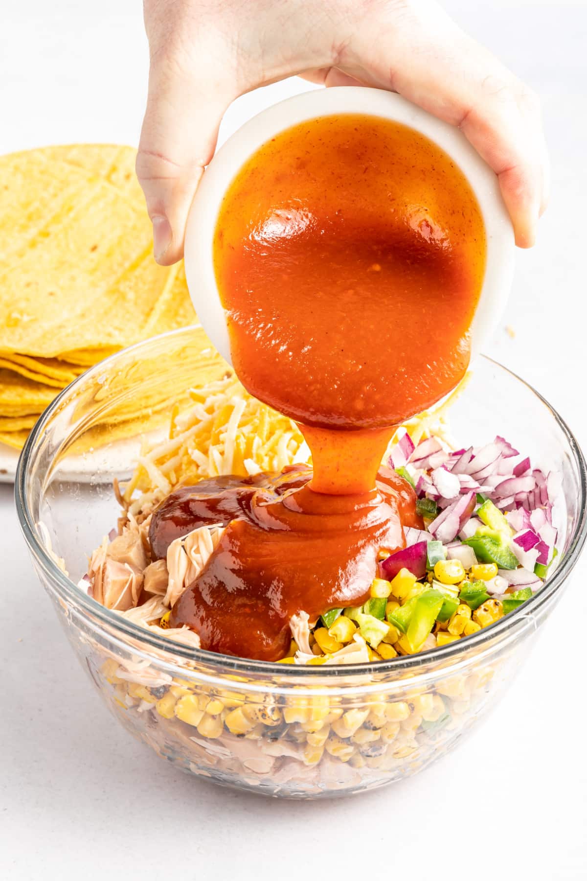 A hand pouring BBQ sauce into a bowl of jackfruit taquito filling. A stack of corn tortillas in background.