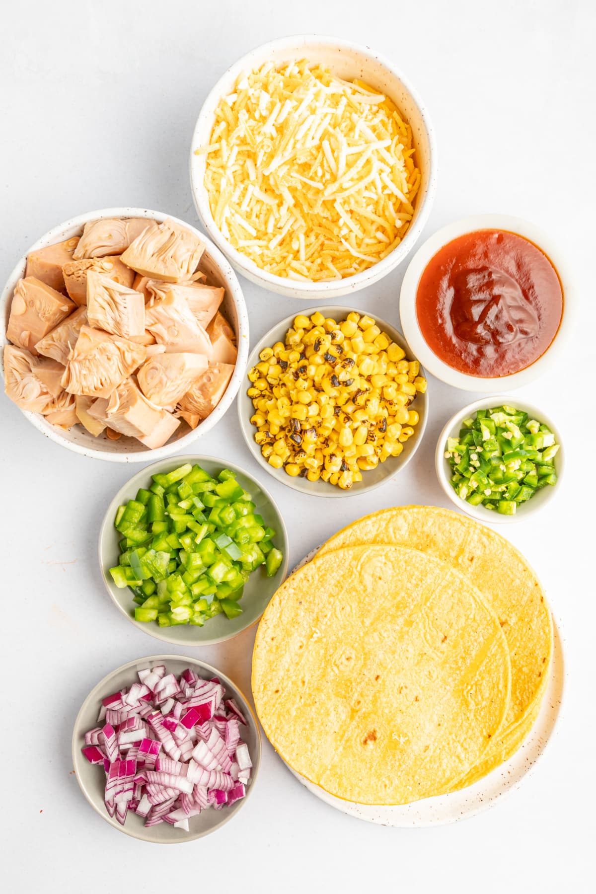 Separate bowls of ingredients for BBQ taquitos: jackfruit, cheese, fire roasted corn, died red onion, diced jalapeno peppers, and BBQ sauce.
