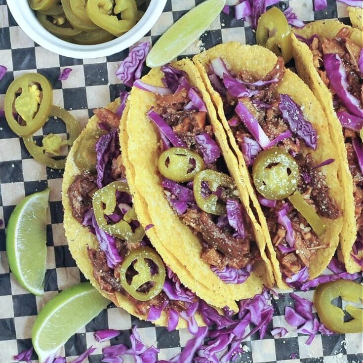 BBQ pulled porcini tacos on black and white checkered paper. Limes, purple cabbage, and sliced jalapeños on the side.