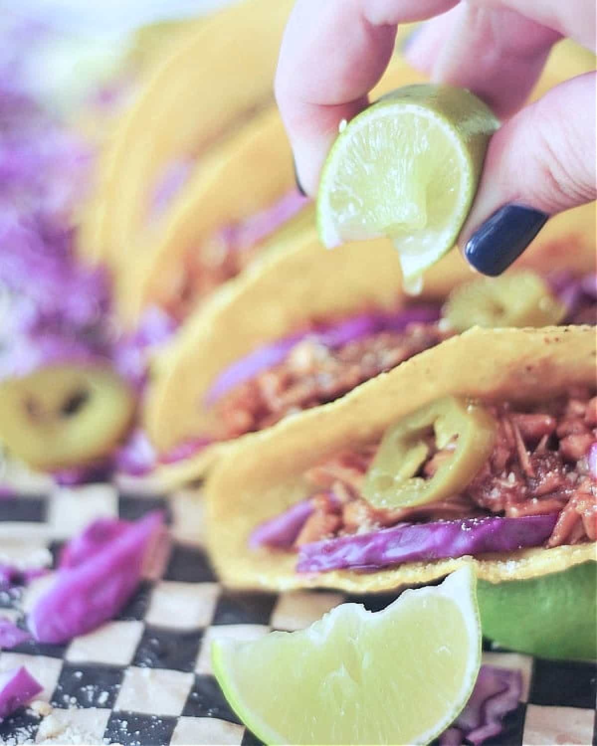 A hand squeezing a lime over BBQ pulled porcini tacos on black and white checkered paper.