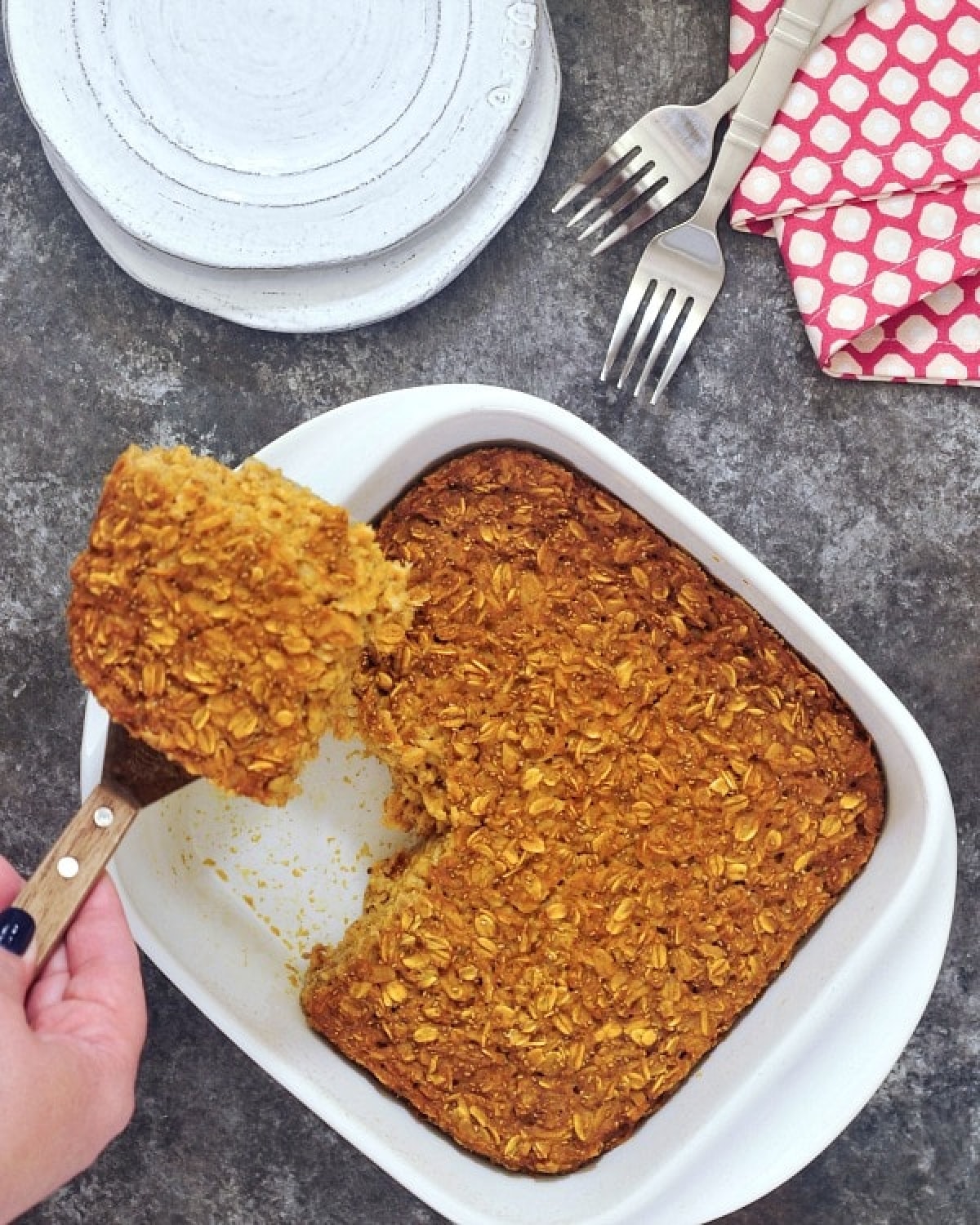 A white ceramic baking pan of baked pumpkin oatmeal, a stack of plates and forks on the side. A hand holding a small spatula is taking one square slice of oatmeal from the baking dish.