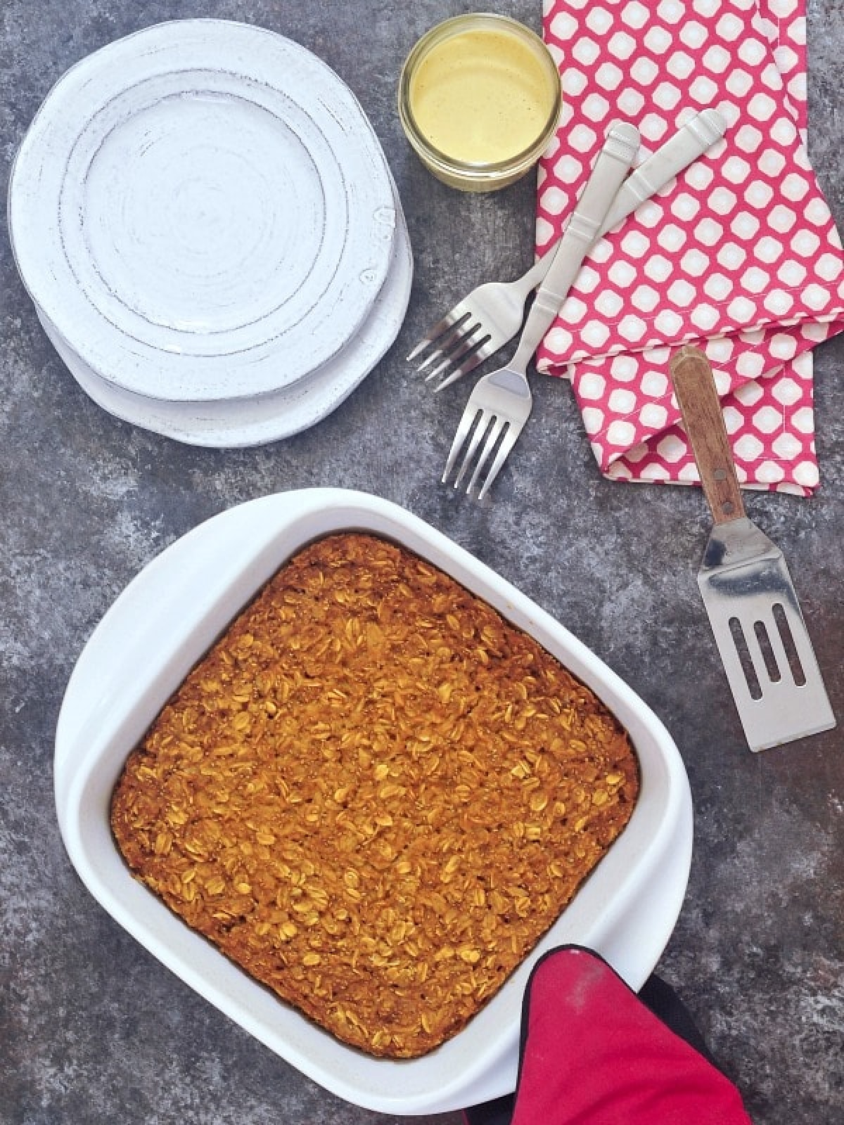 Overhead view of a white ceramic baking pan of baked pumpkin oatmeal held with a red oven glove, with a small glass jar of golden cashew cream, a stack of plates and forks on the side.