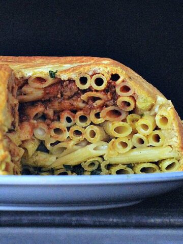 A side view of a large pasta filled pastry dome called a timpano sits on a serving platter with slices cut out to show insides: a red marinara layer of penne pasta on top, a middle layer of white Alfredo pasta, and a bottom layer of green pesto pasta.