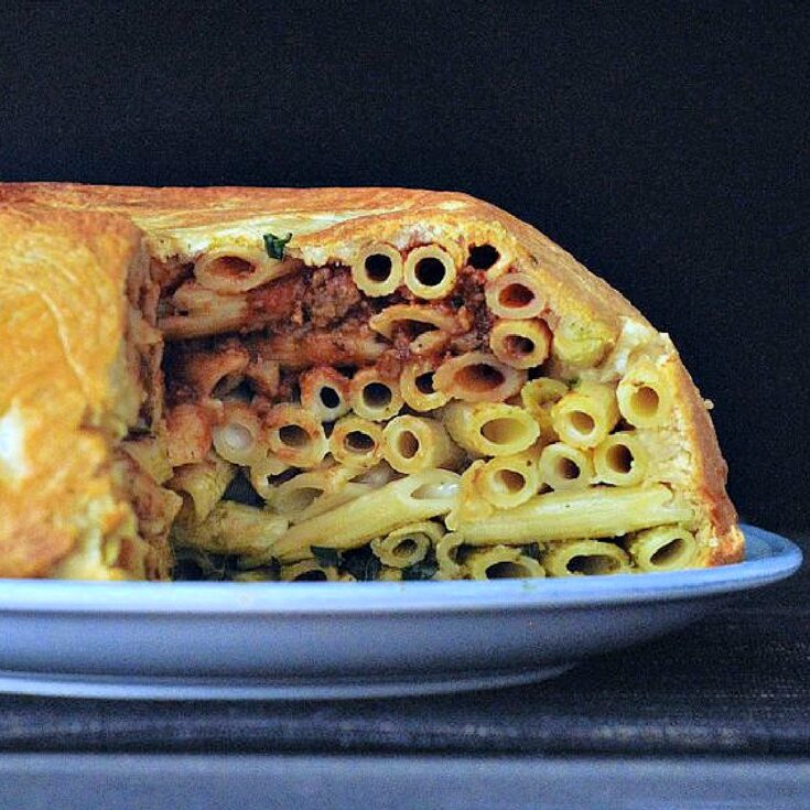 A side view of a large pasta filled pastry dome called a timpano sits on a serving platter with slices cut out to show insides: a red marinara layer of penne pasta on top, a middle layer of white Alfredo pasta, and a bottom layer of green pesto pasta.