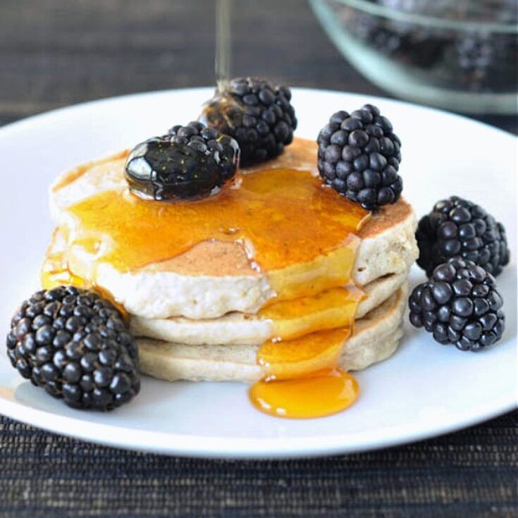 Gluten free buckwheat banana pancakes stacked on a plate with fresh blackberries and syrup being poured over top.