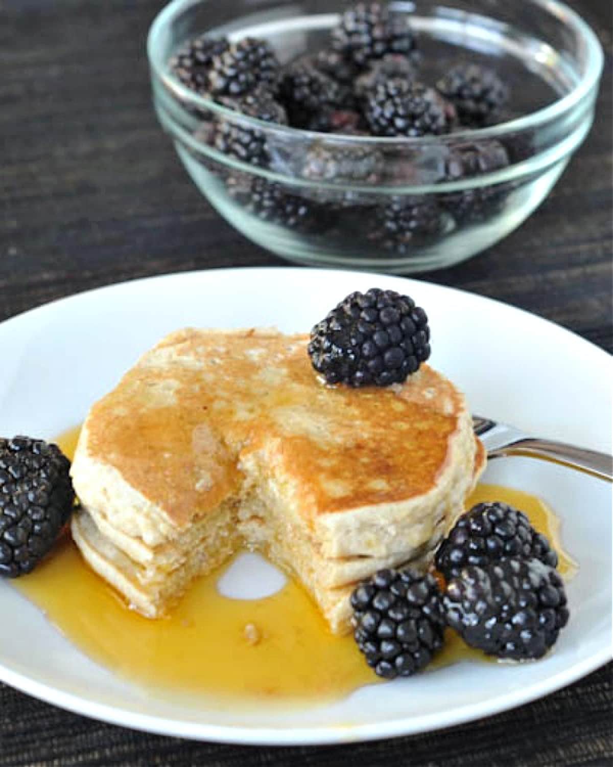 Buckwheat banana pancakes with fresh blackberries and syrup, on a plate with one bite sliced out.