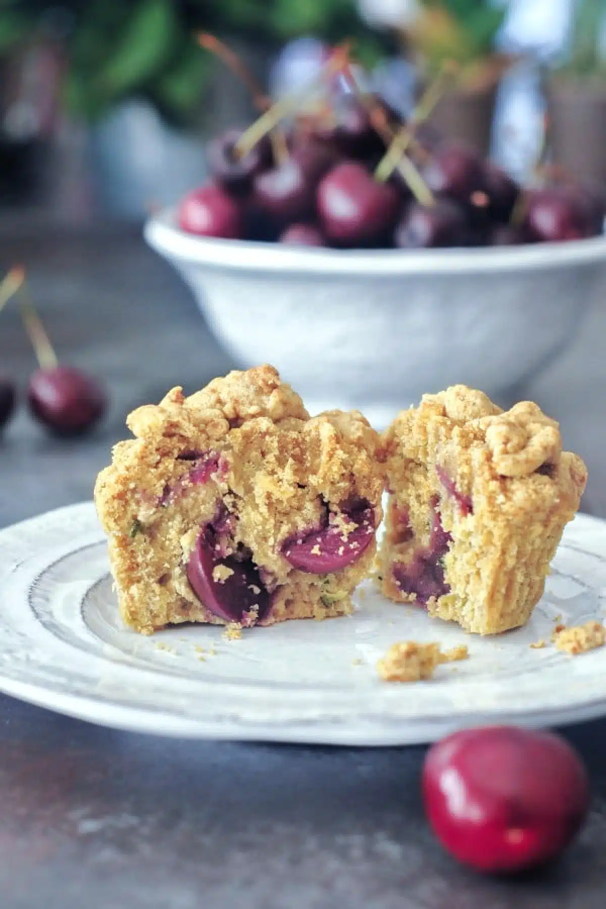 Cherry zucchini muffin sliced in half sitting on a white plate, with a bowl of cherries in background.