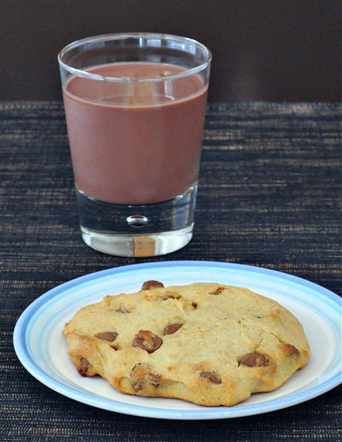A single serve chocolate chip cookie on a white plate. A short glass of chocolate milk sits next to the plate.