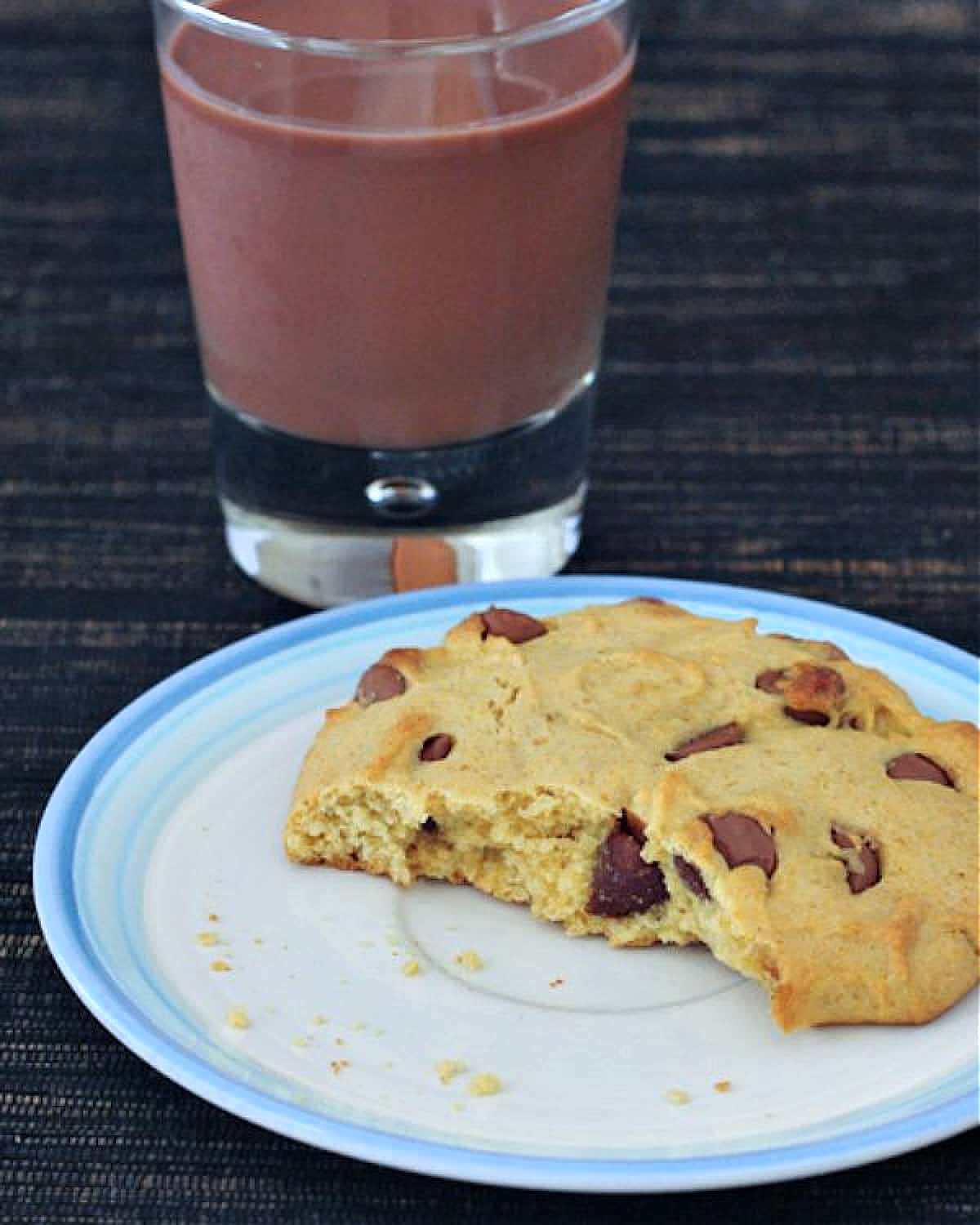 A single serve chocolate chip cookie with a bite out of it on a white plate. A short glass of chocolate milk sits next to the plate.