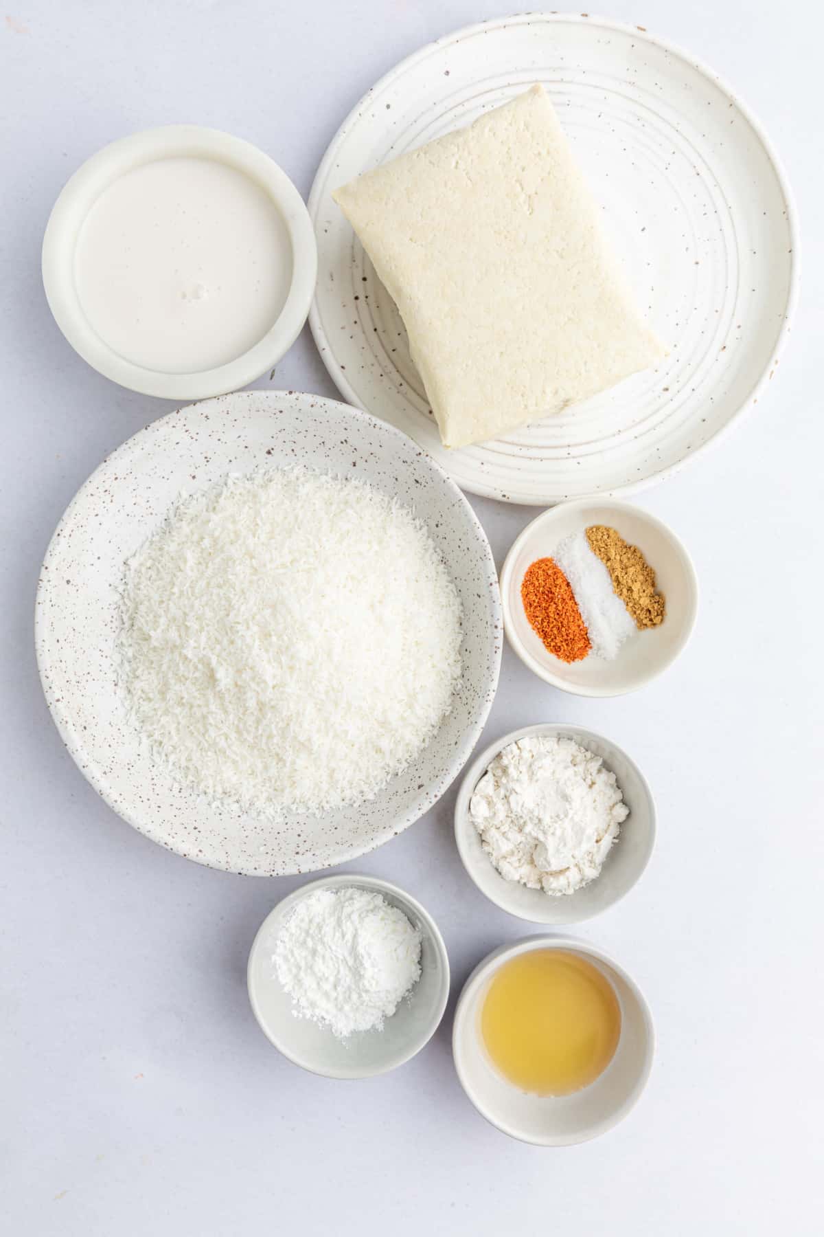 Overhead view of ingredients for crispy coconut tofu in separate bowls: milk, block of tofu, shredded coconut, spices, flour, starch, and maple syrup.