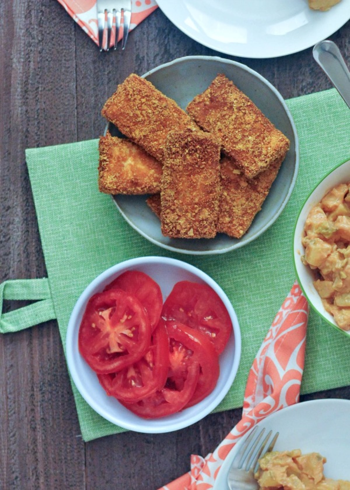 Overhead view of several slices of crispy fried tofu on a grey dish next to a dish of sliced tomatoes and a bowl of potato salad.