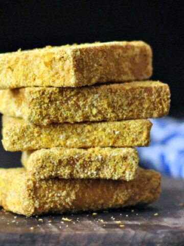 A stack of crispy fried tofu on a dark wood cutting board against a black background, blue and white napkin blurred in the right side background.