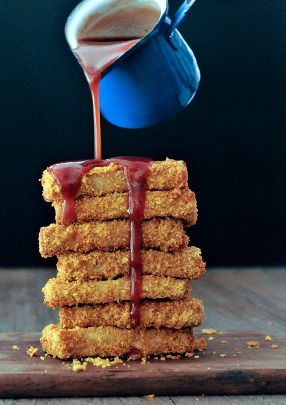 A stack of crispy fried tofu on a dark wood cutting board against a black background, with a blue sauce ladle pouring BBQ sauce over top.