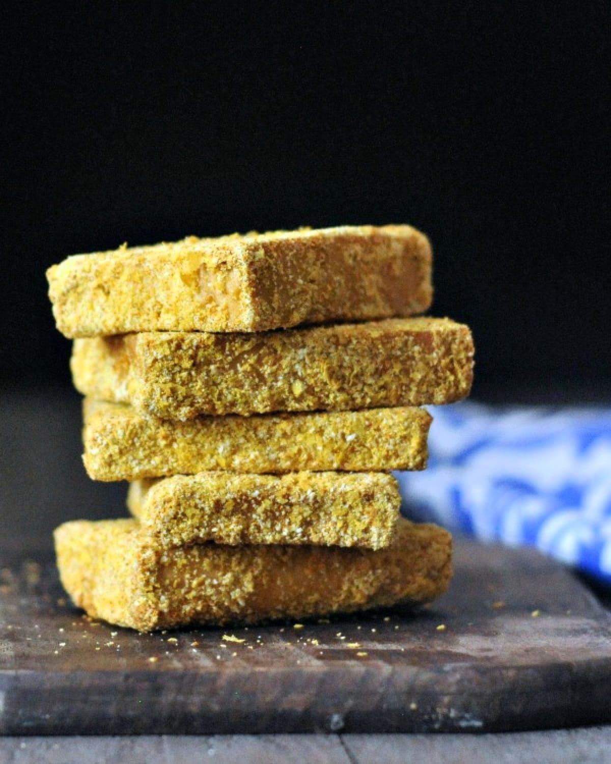A stack of crispy fried tofu on a dark wood cutting board against a black background, blue and white napkin blurred in the right side background.