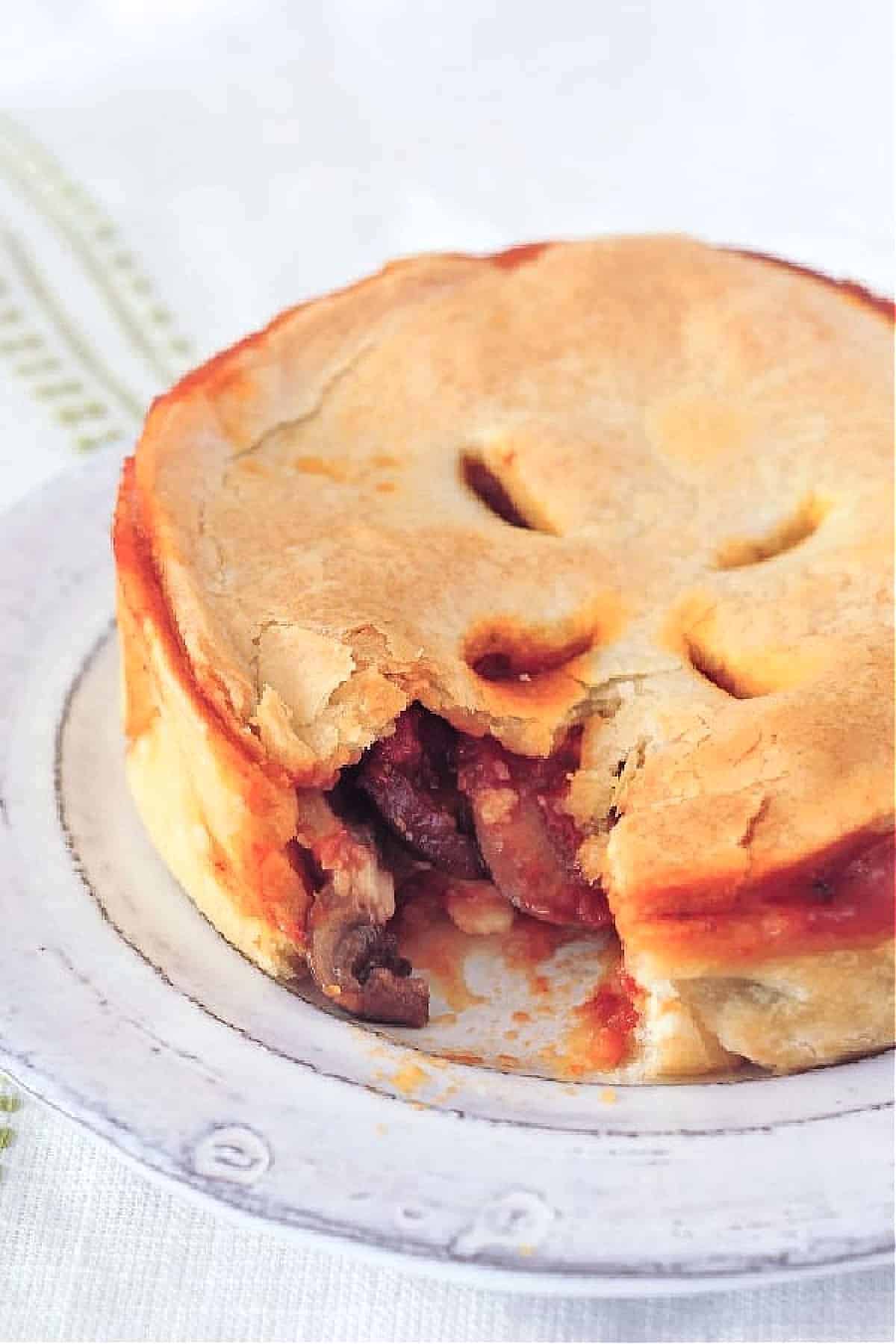 A mini deep dish pizza pot pie on a dish, one bite removed to show filling of mushrooms, sauce, cheese.